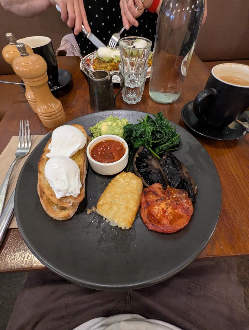 A breakfast plate with poached eggs on toast, a hash brown, grilled tomato, mushrooms, greens, avocado and a pot of red relish