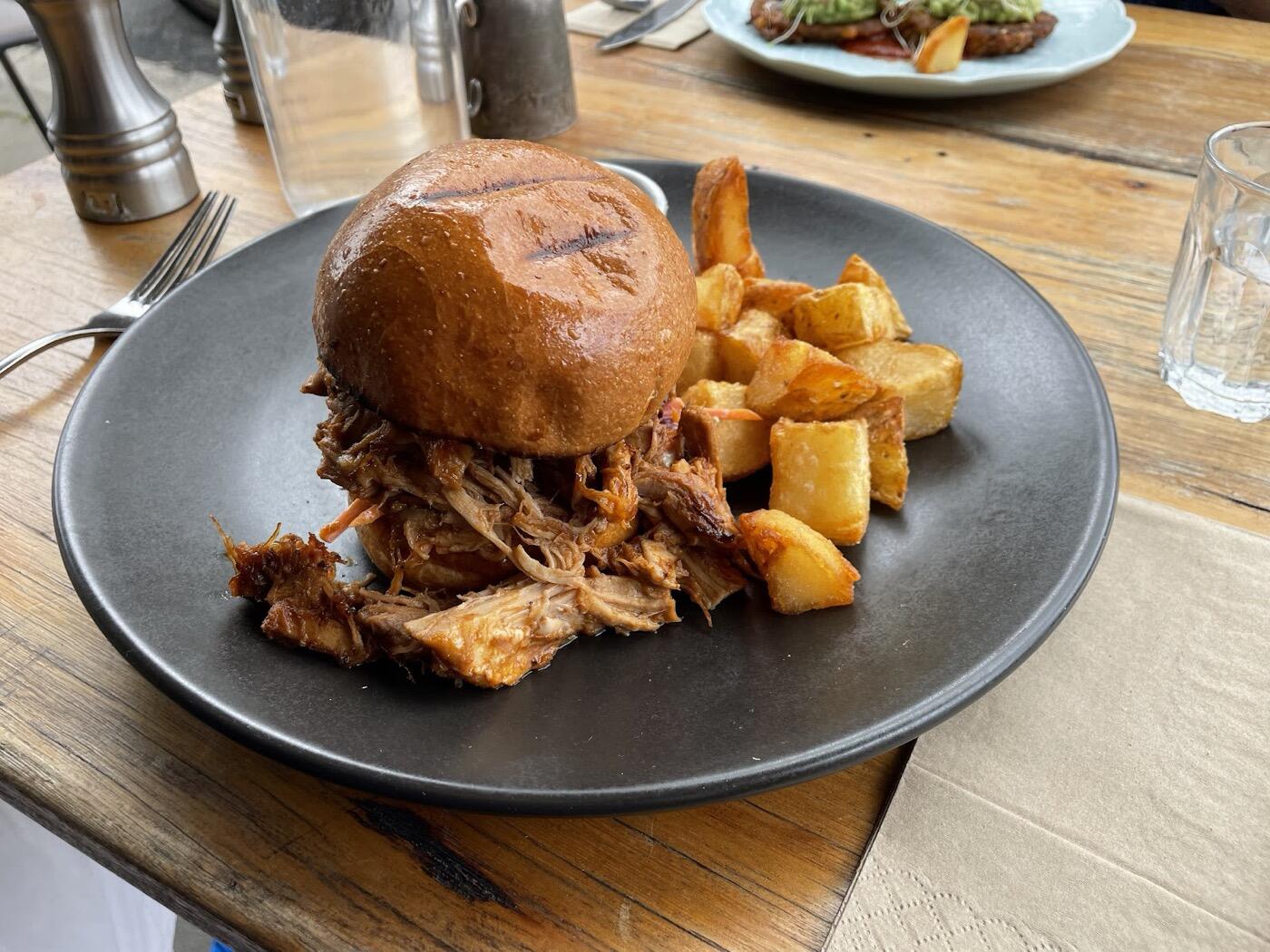 A burger with a glossy bun and a pulled-meat filling beside golden potato chunks on a dark plate