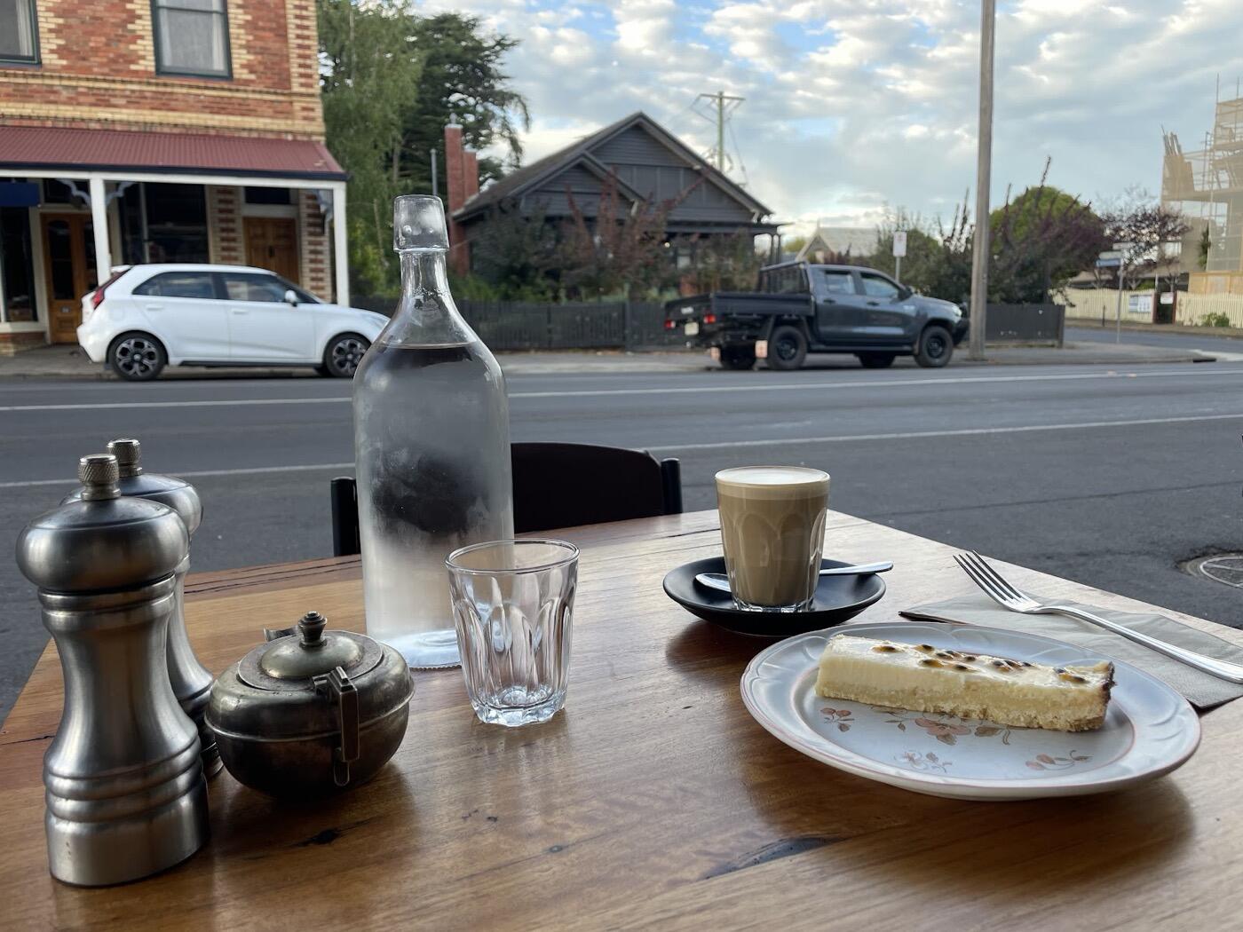 A slice of cake and a small coffee on a footpath table, next to a water bottle and a pepper grinder, with the street behind