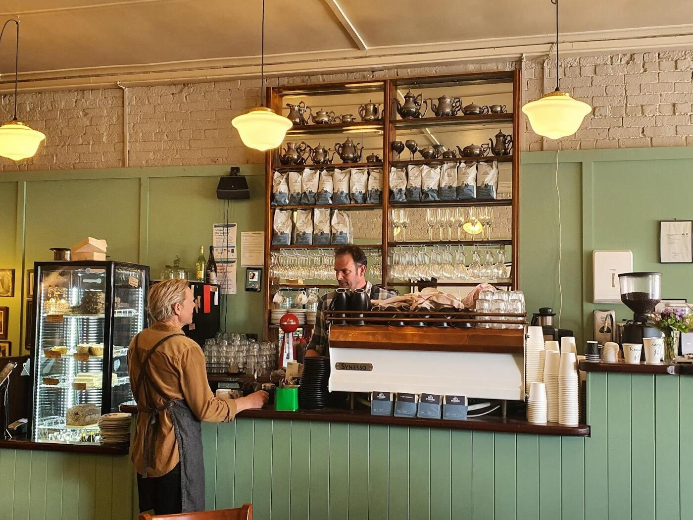 The café counter with a green panelled front and an espresso machine, with shelves of teapots and coffee bags behind