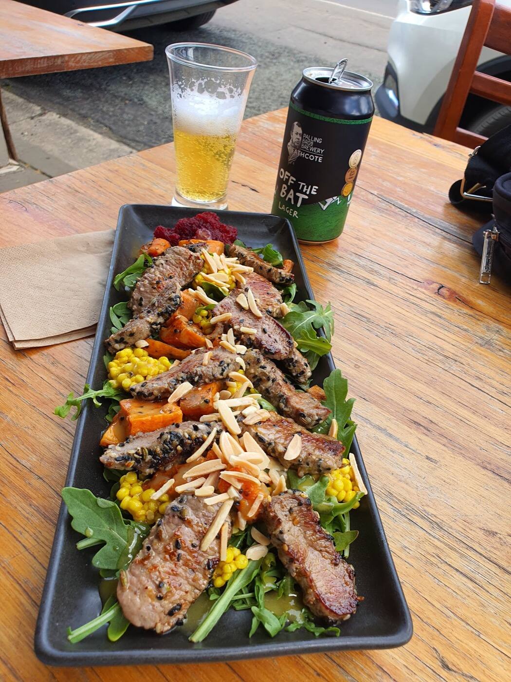 A long dark plate of sliced herb-crusted meat on rocket and roasted pumpkin with flaked almonds, beside a glass of beer