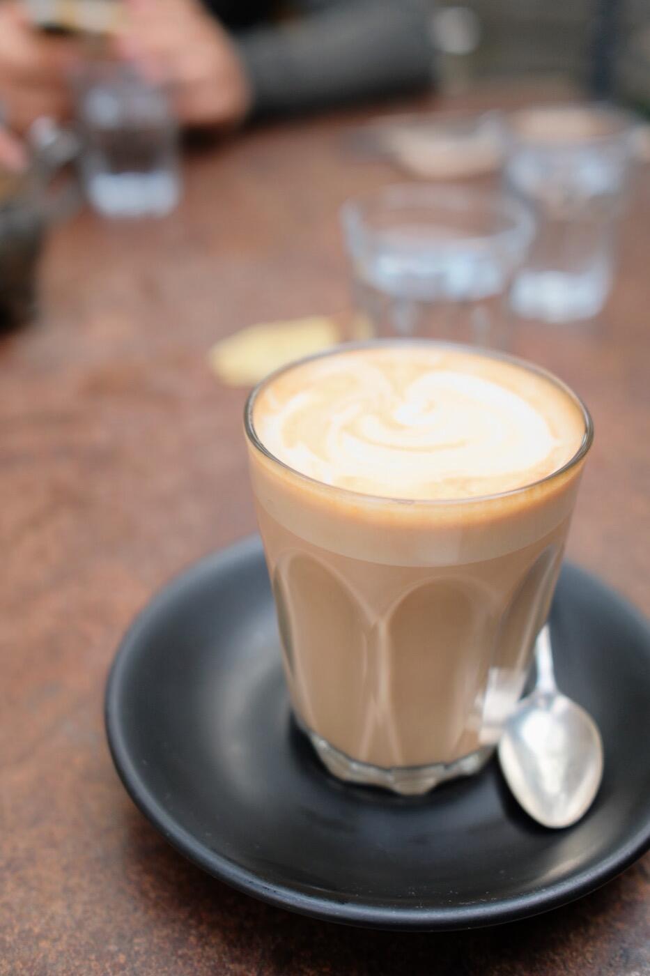 A latte in a glass on a black saucer with a spoon, on a timber table