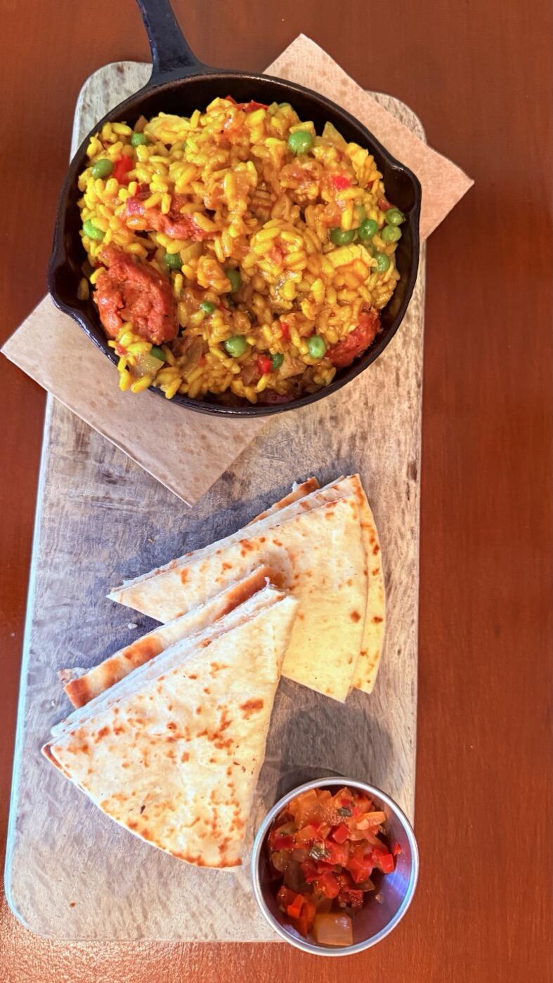 A small black skillet of saffron-yellow paella with chorizo and peas, on a board with flatbread triangles and a pot of red salsa, seen from above