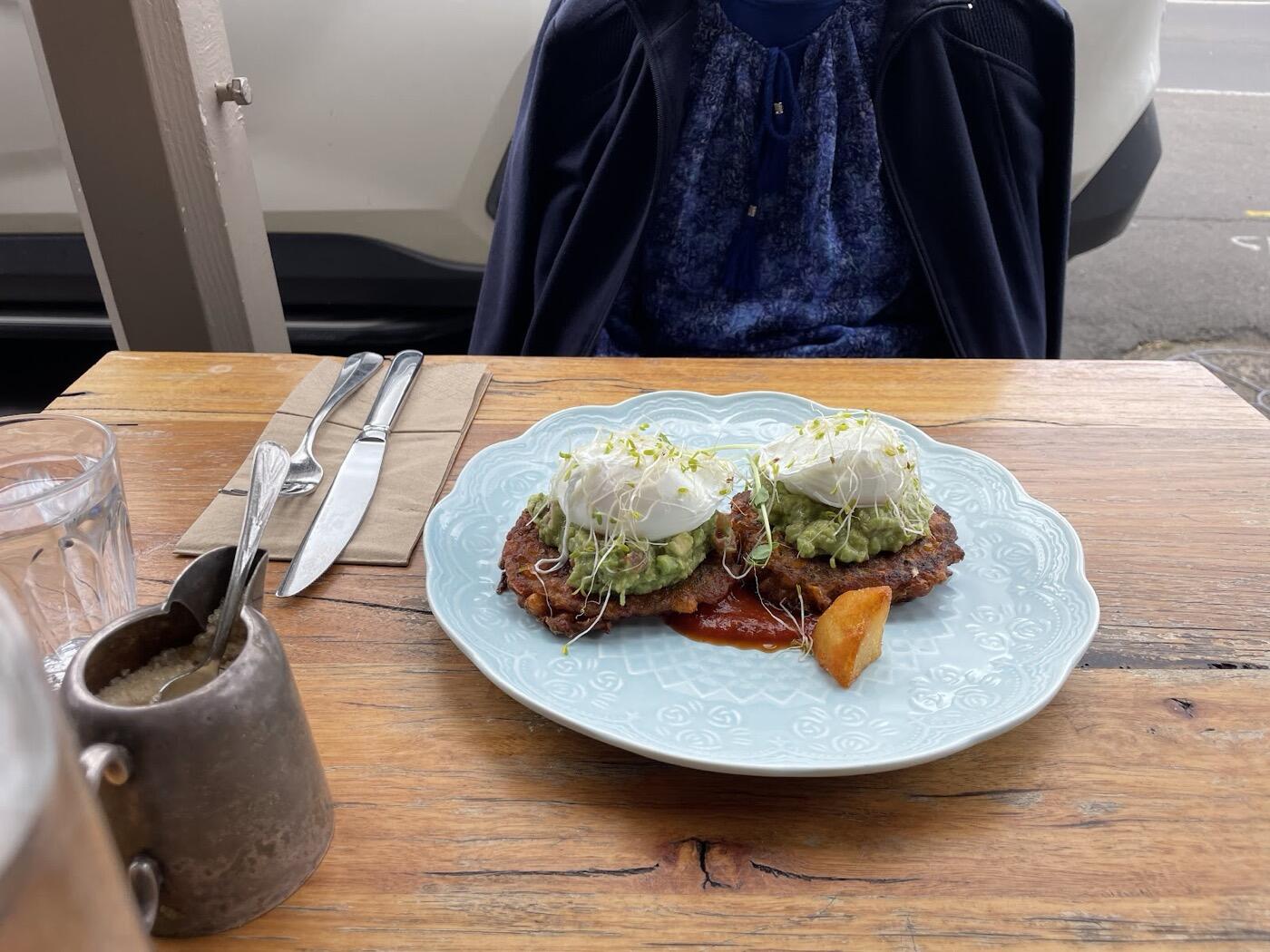 Two poached eggs with smashed avocado and sprouts on a pale blue scalloped plate, with a roast potato wedge